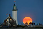 A full harvest moon rises over the ocean as Scituate Lighthouse rises in the foreground, as a crowd stood on the breakwater next to it to watch it.