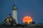 A full harvest moon rises over the ocean as Scituate Lighthouse rises in the foreground, as a crowd stood on the breakwater next to it to watch it.