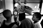 A boxing program called Bonding in Brotherhood was started at Holmes Elementary School, gathering kids most in need of calm and focus. The students were struggling. Some were withdrawn. Some were dealing with domestic violence and the death of a parent. Some were written up constantly for aggressive behavior. The students gather twice a week, and before they box, have a stretching and warmup session. They still have struggles, but nothing like before.
Boxing coach Russell Kimber carries a box of boxing gloves down to the school gym with student Keith Evans, age 8, by his side.